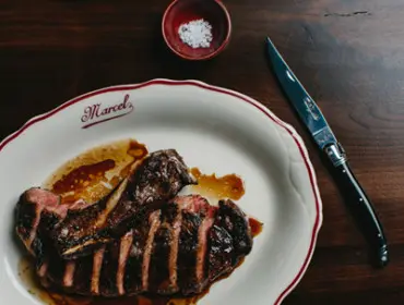 Sliced steak with sauce on a white plate with a red rim, next to a Laguiole knife and a small dish of salt.