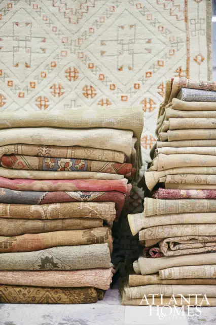 Stacks of folded rugs in neutral tones with geometric patterns and pops of red and pink. A large rug with a cream background and orange geometric design is visible behind the stacks.