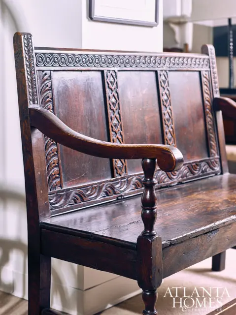 Dark wood antique bench with carved detailing on the backrest and arms, featuring a turned spindle on the armrest support.