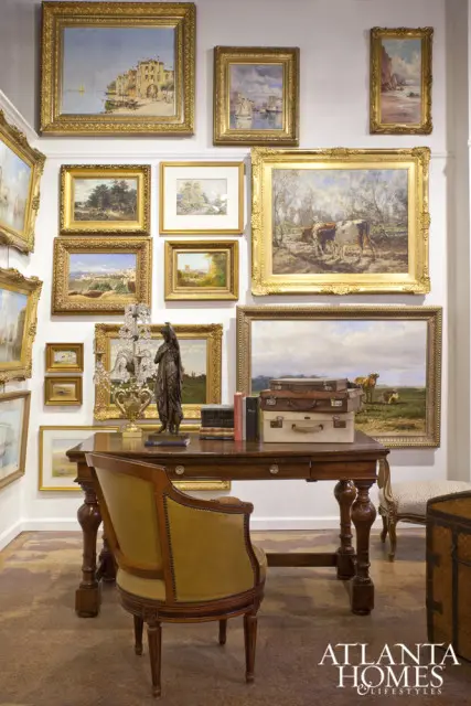 Desk with stacked suitcases and books, surrounded by framed paintings of landscapes and seascapes, with a leather armchair in the foreground.