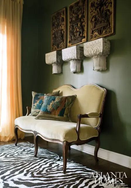Green walls with three ornate gold wall panels and three white carved corbels above a light yellow velvet settee with patterned pillows on a zebra print rug.