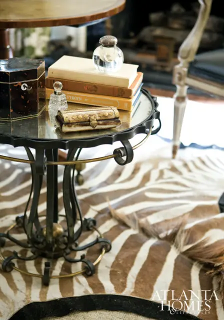 Round mirrored-top side table with ornate metal base, stacked books, decorative boxes, and a zebra print rug.