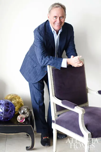 Man in a blue plaid suit leaning on a purple velvet armchair, with decorative orbs on a black table to his left.