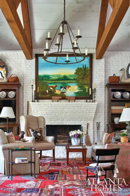 Living room with white brick fireplace, large pastoral painting, wingback chairs, and a red oriental rug. Exposed wooden beams accent the ceiling.