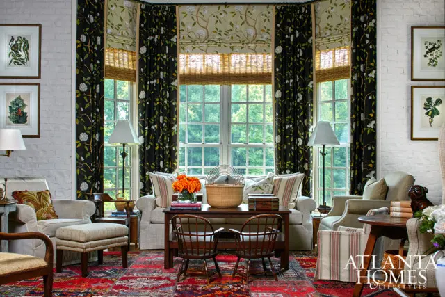 Living room with white brick walls, bay windows with floral curtains and bamboo blinds, upholstered chairs, wooden furniture, and a red Persian rug.