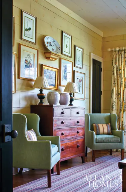 Living room with light wood paneled walls, two green wingback chairs, a wooden dresser with lamps and vases, and botanical prints on the wall.