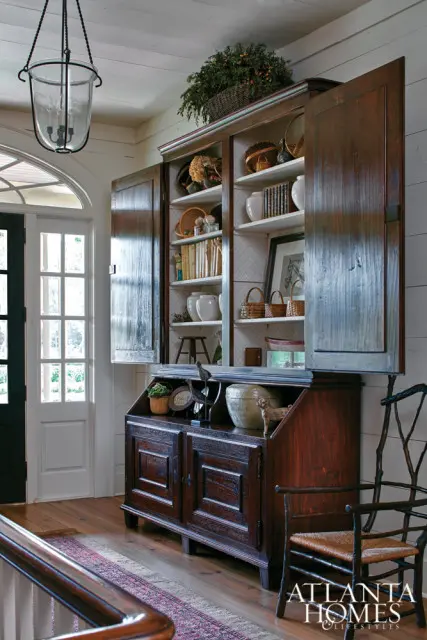 Dark wood cabinet with open doors displaying books, pottery, and baskets. A glass pendant light hangs overhead, and a woven chair sits to the right.