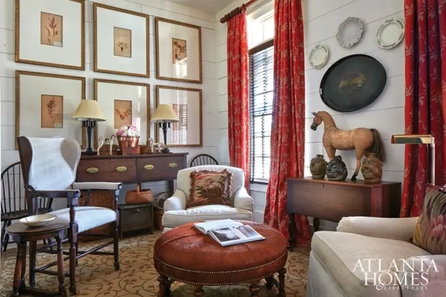 Living room with white shiplap walls, red floral curtains, and a collection of framed botanical prints. Furniture includes a wingback chair, an upholstered ottoman, and a sofa.