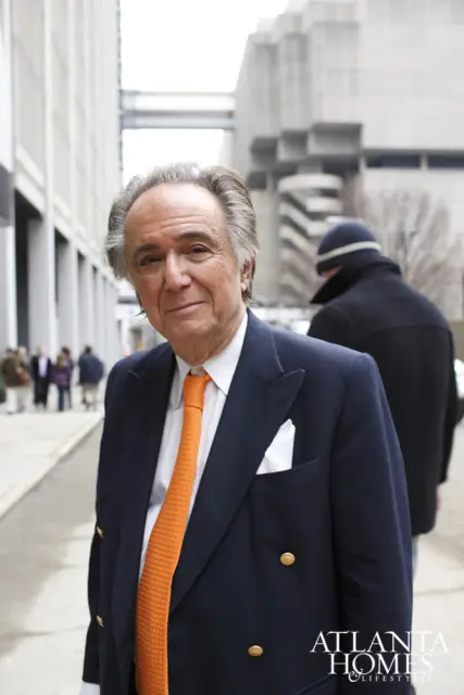 Man in a navy blue suit with an orange tie and white pocket square, standing outside with buildings in the background.