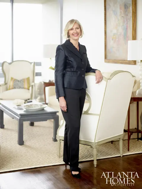 Woman in a black suit standing in a living room with a painting on the wall and a white chair.