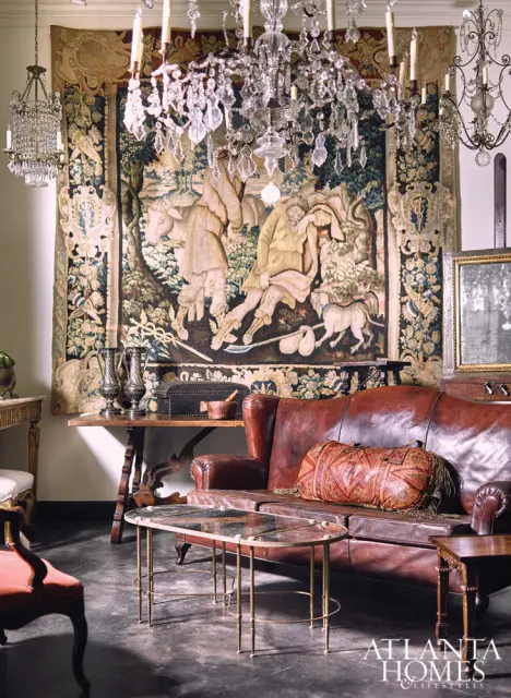 Brown leather sofa with patterned lumbar pillow in front of a large tapestry and crystal chandeliers. A glass and brass coffee table sits in front of the sofa.