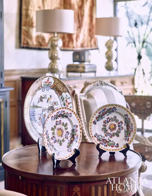 Three decorative plates with floral and bird motifs displayed on a wooden table in a room with lamps and ornate furniture.