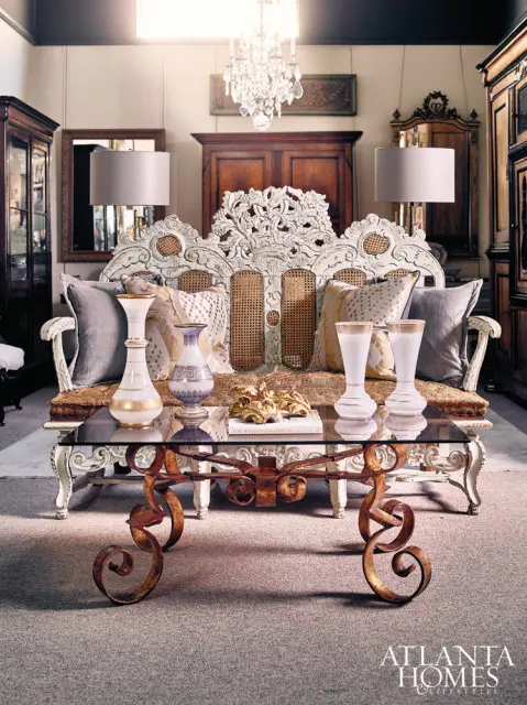 Living room with ornate white carved sofa and cane back, glass-top coffee table with scrolled metal base, crystal chandelier, and antique cabinets.