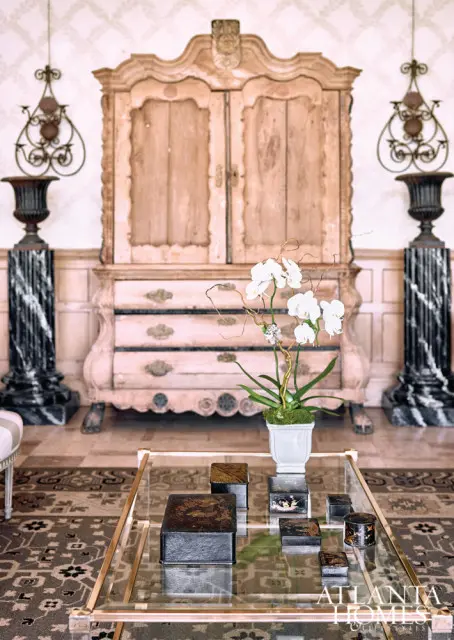 Living room with antique wooden armoire, marble pedestals with urns, and a glass coffee table displaying decorative boxes and a white orchid plant.