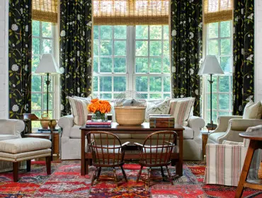 Living room with white brick walls, floral curtains, upholstered sofas and armchairs, a wooden coffee table with a basket and flowers, and a colorful oriental rug.