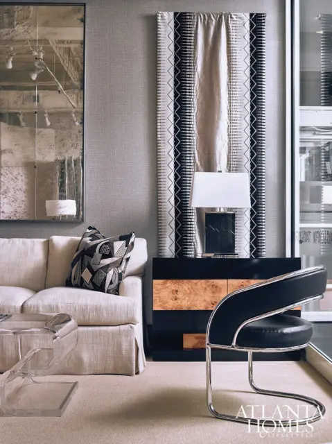 Living room with a cream sofa, abstract patterned pillow, burl wood and black dresser, and a black leather chair with chrome accents. A large distressed mirror hangs on the wall.