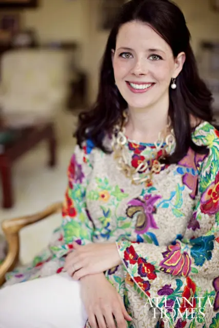 Woman with dark hair and green eyes smiling, wearing a colorful floral print top and a chunky gold necklace.