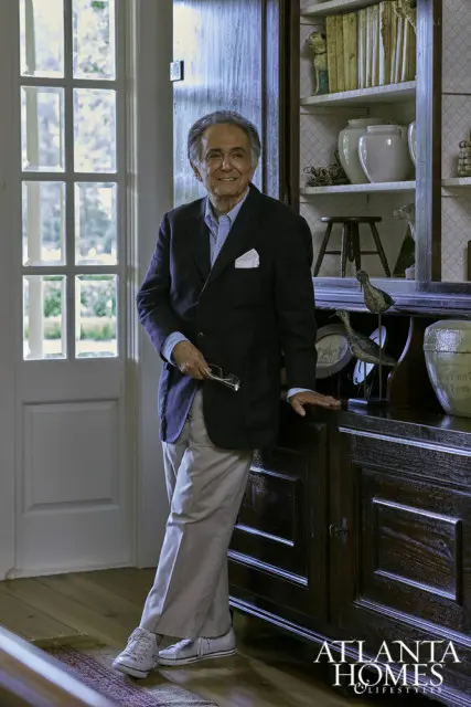Man in a navy blazer, light blue shirt, and tan pants holding glasses, standing next to a dark wood cabinet filled with books and decorative objects. Atlanta Homes & Lifestyles logo in the lower right corner.