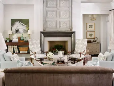 Living room with a fireplace topped by antique doors, flanked by patterned armchairs and light blue gingham sofas. Two teal lamps with pleated shades sit on side tables.
