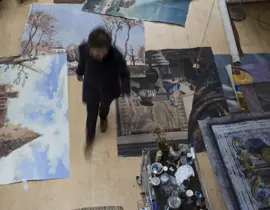 Overhead view of an artist walking through a studio filled with paintings and art supplies.