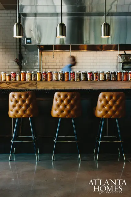 Brown tufted bar stools in front of a counter displaying rows of glass jars filled with colorful ingredients. Stainless steel range hood and subway tile backsplash in background.