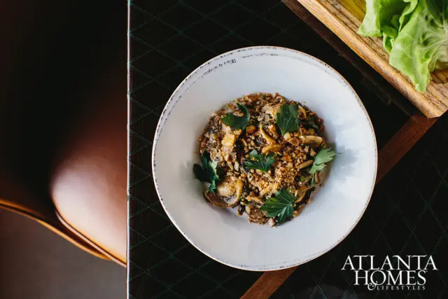 White bowl of mushroom and nut dish garnished with parsley, next to lettuce leaves on a wooden board. Atlanta Homes & Lifestyles logo in the corner.