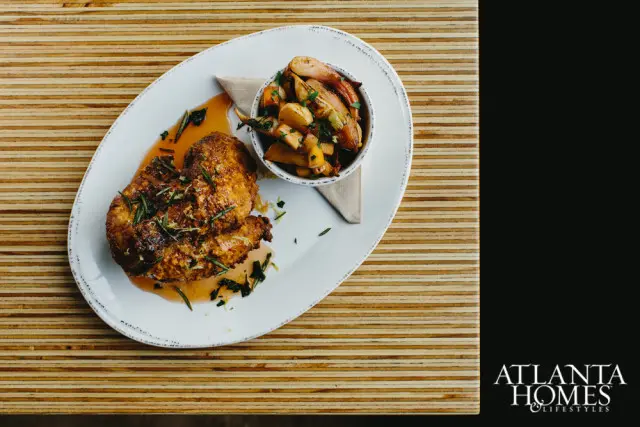 Roasted chicken with herbs and lemon zest served with roasted root vegetables on a white oval plate, set on a striped wooden surface. Atlanta Homes & Lifestyles logo in the bottom right corner.