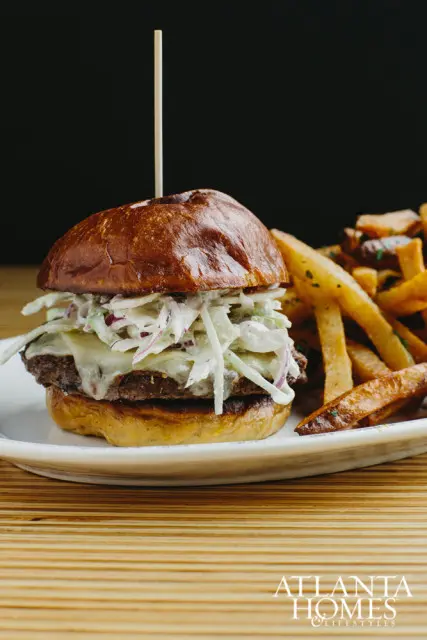 Burger with coleslaw and melted cheese, served with french fries on a white plate with a wooden background.