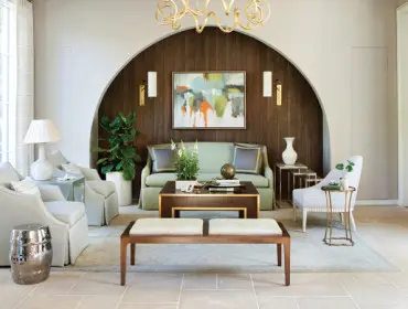 Living room with light gray upholstered chairs, a pale green sofa, and a dark wood paneled arched wall featuring abstract art and gold sconces.