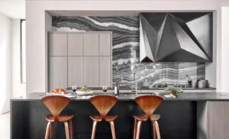 Modern kitchen with three wood counter stools in front of a dark countertop island. The backsplash features dramatic black and white veined stone and a geometric metal range hood.