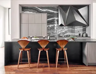 Modern kitchen with three wood counter stools in front of a dark countertop island. The backsplash features dramatic black and white veined stone and a geometric metal range hood.