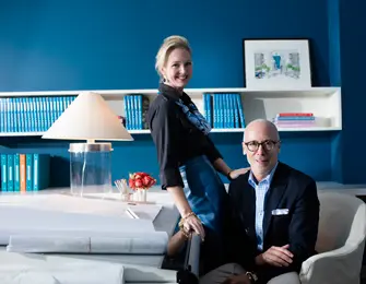 A woman in a black shirt and blue skirt leans against a desk, while a man in a blue blazer and glasses sits in a chair. They are in a room with a bright blue wall and built-in bookshelves.