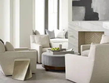 Living room with four upholstered chairs, a gray ottoman with a tray of green apples, a marble fireplace with herringbone tile, and abstract art above the mantel.