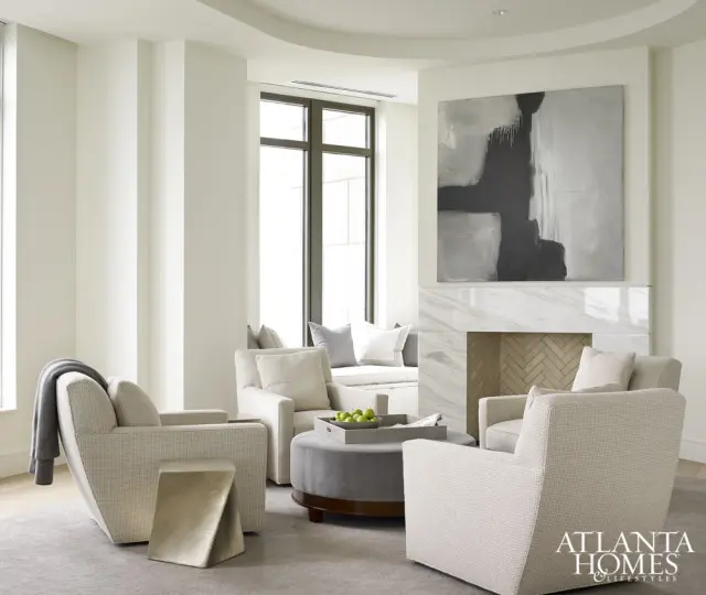 Living room with four upholstered chairs, a gray ottoman with a tray of green apples, a marble fireplace with herringbone tile, and abstract art above the mantel.