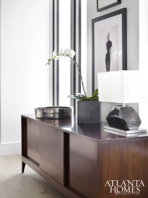 Dark wood credenza with sliding doors topped with a metallic bowl, potted orchid, and a faceted table lamp. Framed art hangs on the wall above.