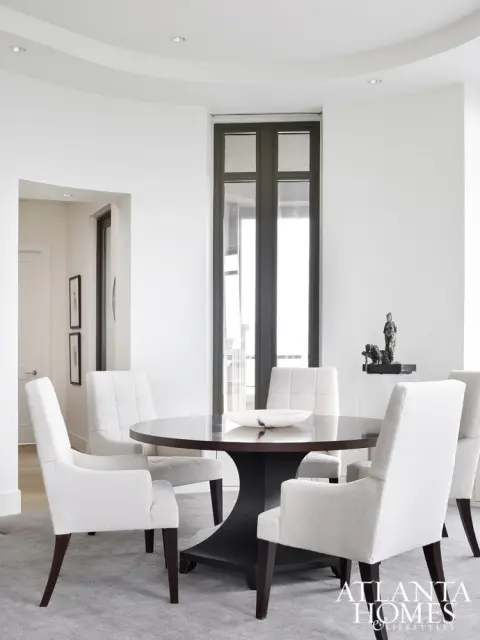 Dining room with dark wood round table, upholstered chairs in cream fabric, and a tall window with dark frame.
