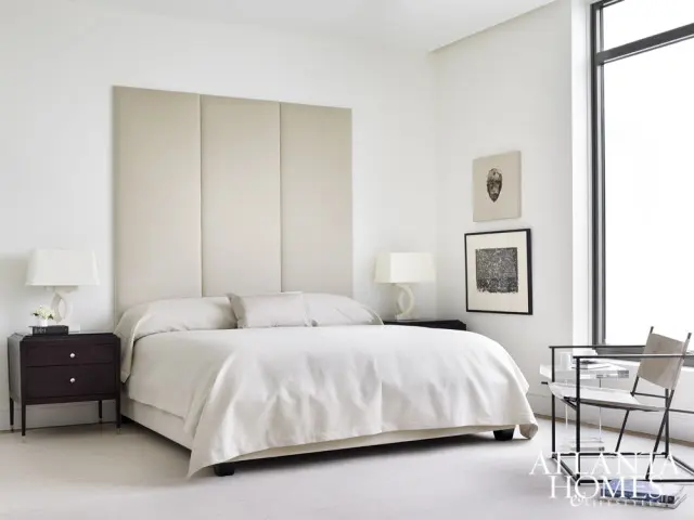 Bedroom with a large upholstered headboard, neutral bedding, dark wood nightstand with a lamp, and modern artwork on the wall.