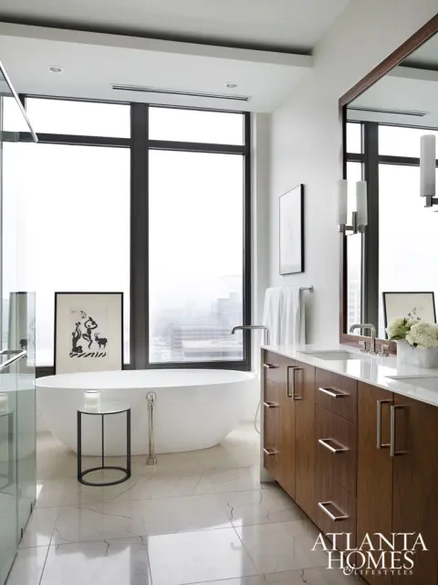 Modern bathroom with freestanding white bathtub, marble tile floors, and a long wooden vanity with dual sinks and chrome hardware. Large windows offer city views.