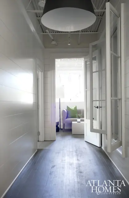 Hallway with white shiplap walls, dark wood floors, and a large black pendant light. A doorway opens to a sitting area with a lavender sofa, green pillow, and white coffee table.