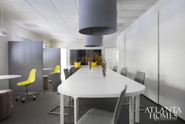 Modern conference room with a long white table, gray upholstered chairs, yellow accent pillows, and large gray pendant lights overhead.