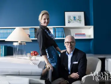 A woman in a black shirt and blue skirt leans on a desk next to a man in a navy blazer and plaid shirt. They are in front of a bright blue wall with bookshelves.
