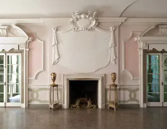Elegant living room with a white ornate fireplace, flanked by two gold urns on pedestals. Light pink walls with decorative molding and two French doors.