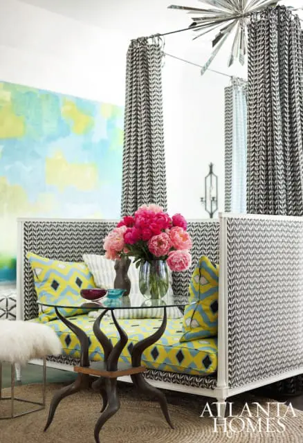 Daybed with yellow and blue geometric print cushions and black and white patterned upholstery, glass top table with sculptural base, and a vase of pink peonies.