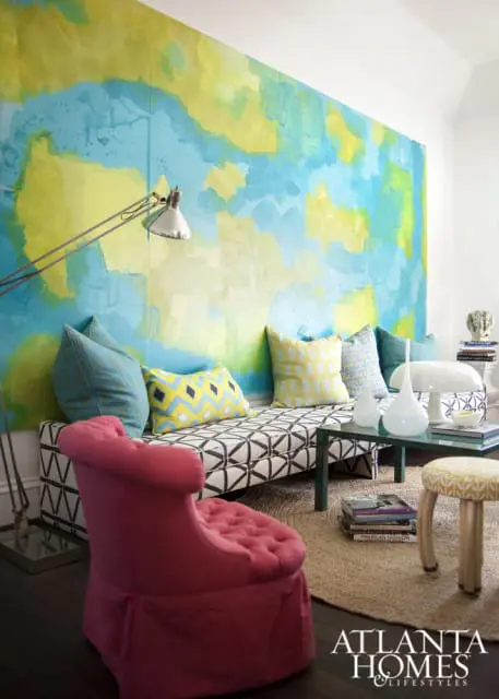 Living room with a large abstract mural in blue and yellow on the wall, a patterned daybed with colorful pillows, a pink tufted armchair, and a glass coffee table.