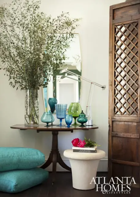 Entryway console table with a large glass vase of greenery, assorted blue and green glass vases, a modern desk lamp, and a decorative wooden screen.