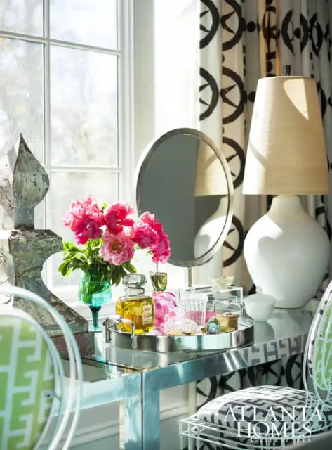 Vanity table with mirrored surface, silver tray holding perfume bottles and pink peonies in a teal vase. White lamp and patterned curtains in background.