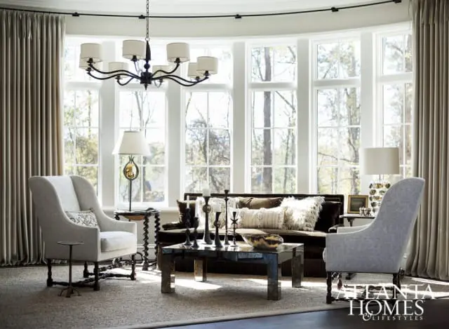 Living room with large bay windows overlooking trees, featuring two upholstered armchairs, a dark sofa with textured pillows, a mirrored coffee table, and a black chandelier.