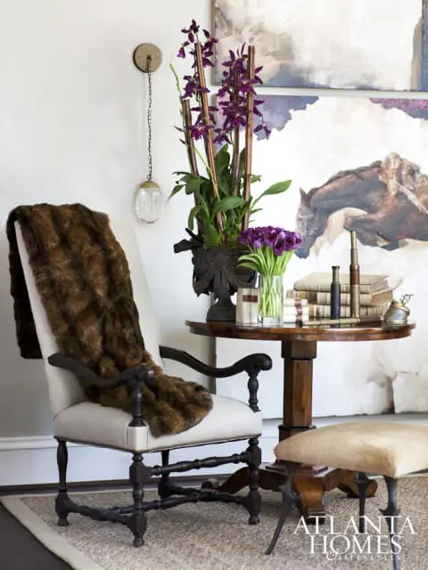 Cream upholstered armchair with dark wood frame draped with a brown fur throw, next to a round wooden table with books, flowers, and a telescope.