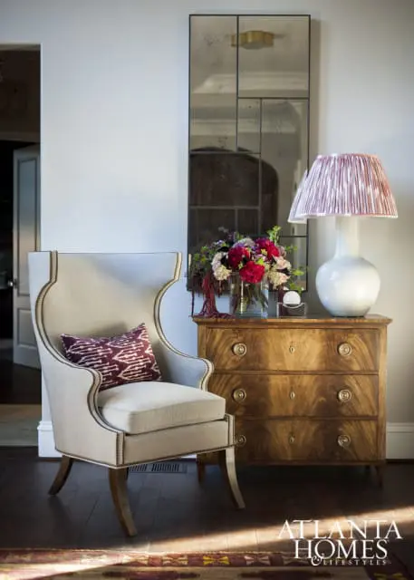 Wingback chair with patterned pillow next to a wooden dresser topped with a lamp and floral arrangement, beneath a tall, segmented mirror.