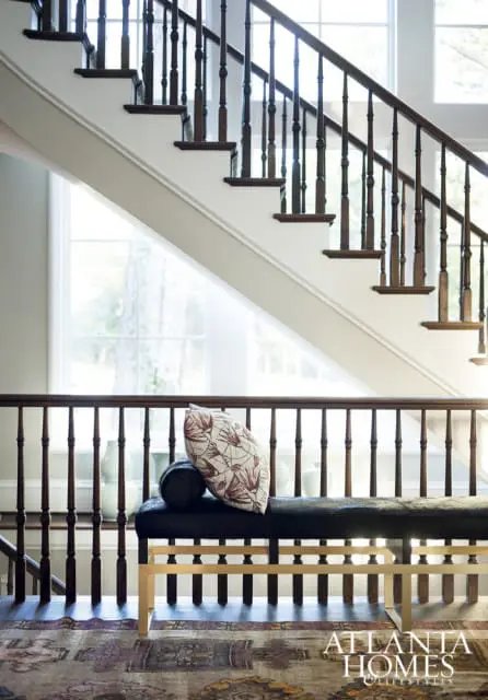 Interior view of a grand staircase with dark wood banister and white risers, a dark upholstered bench with a patterned pillow, and an ornate rug on the floor.